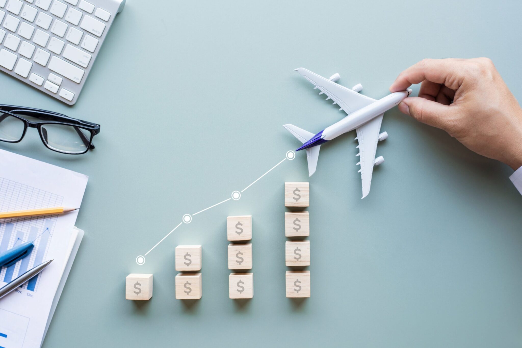 A hand holds a toy airplane above rising stacks of dollar-marked blocks, resembling a growth graph, symbolizing improved airport performance—next to 