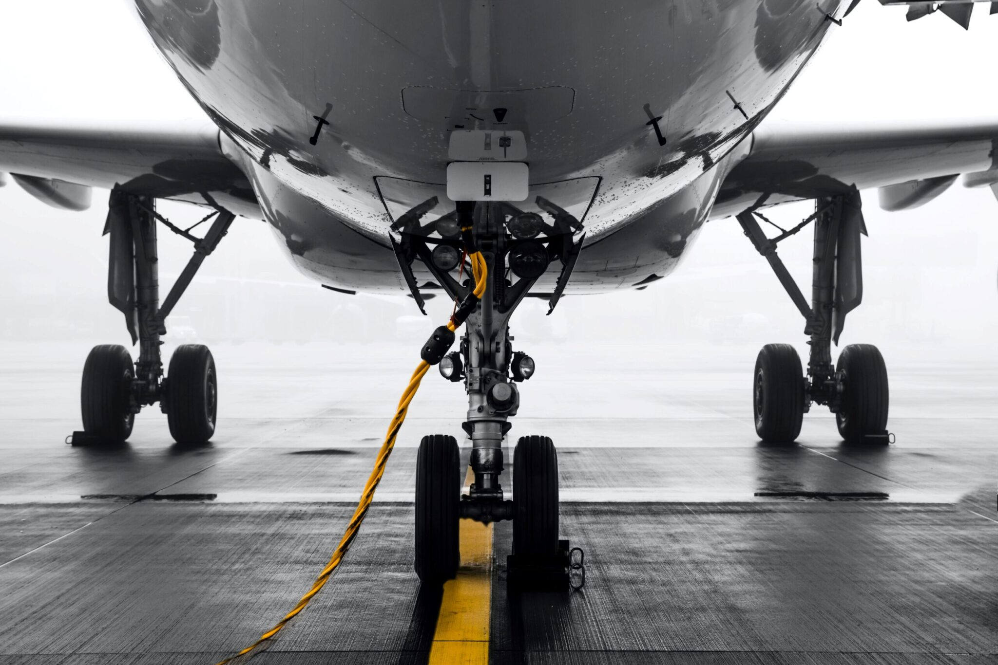 Close-up view of an airplane landing gear with a yellow cable on the runway, mist in the background. Image used for the 