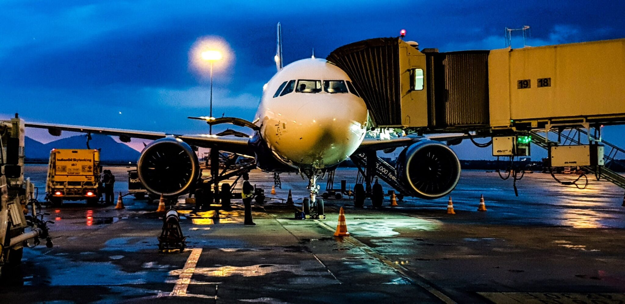 At night, ACRIS Best Practice enhances airport operations as a passenger airplane is parked at the gate with a jet bridge connected, wet tarmac reflecting lights, service vehicles nearby, and a dark blue evening sky overhead.