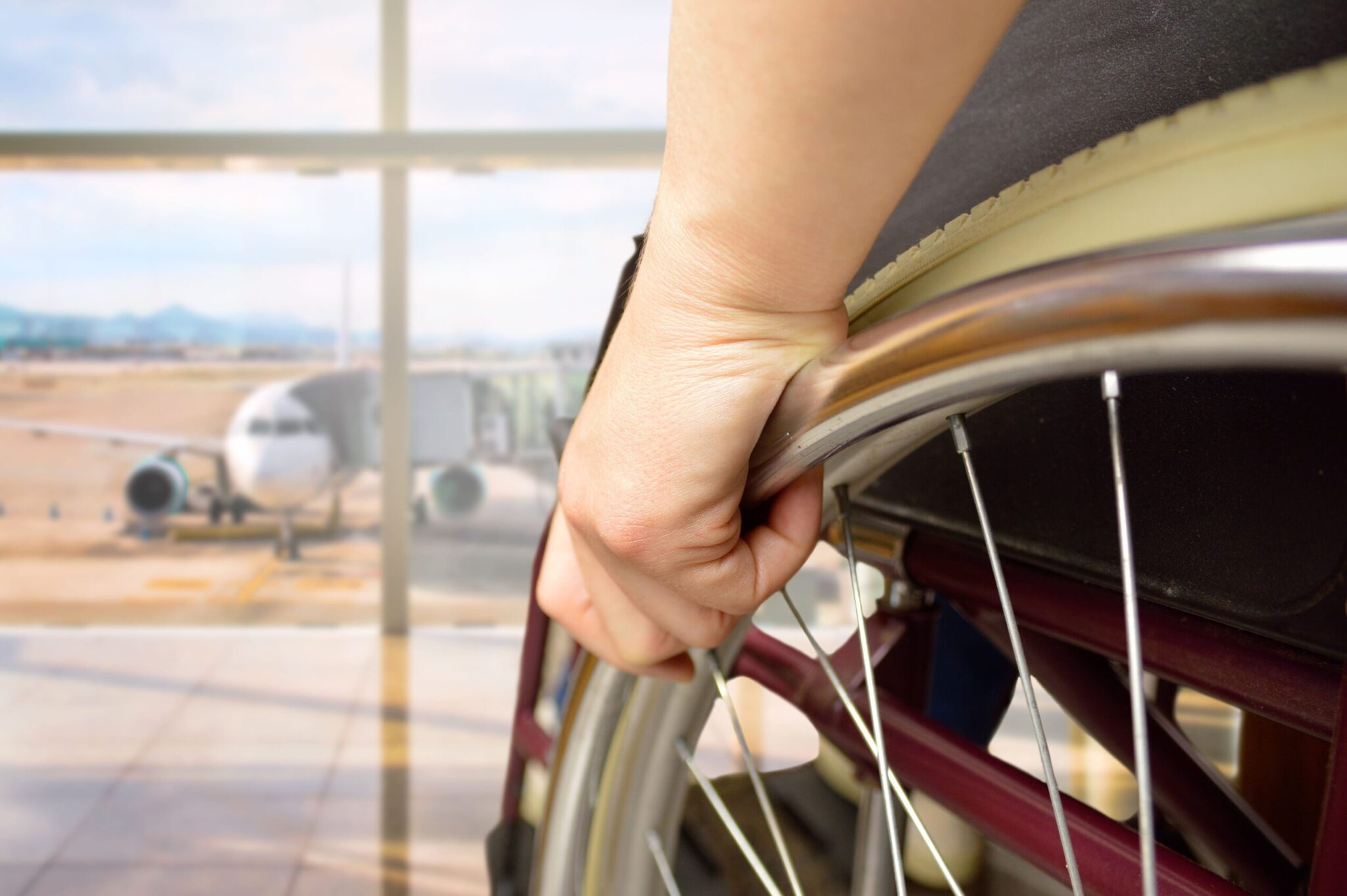 A close-up of a hand on a wheelchair wheel at an airport, with a plane visible outside, illustrates accessible travel as detailed in the 