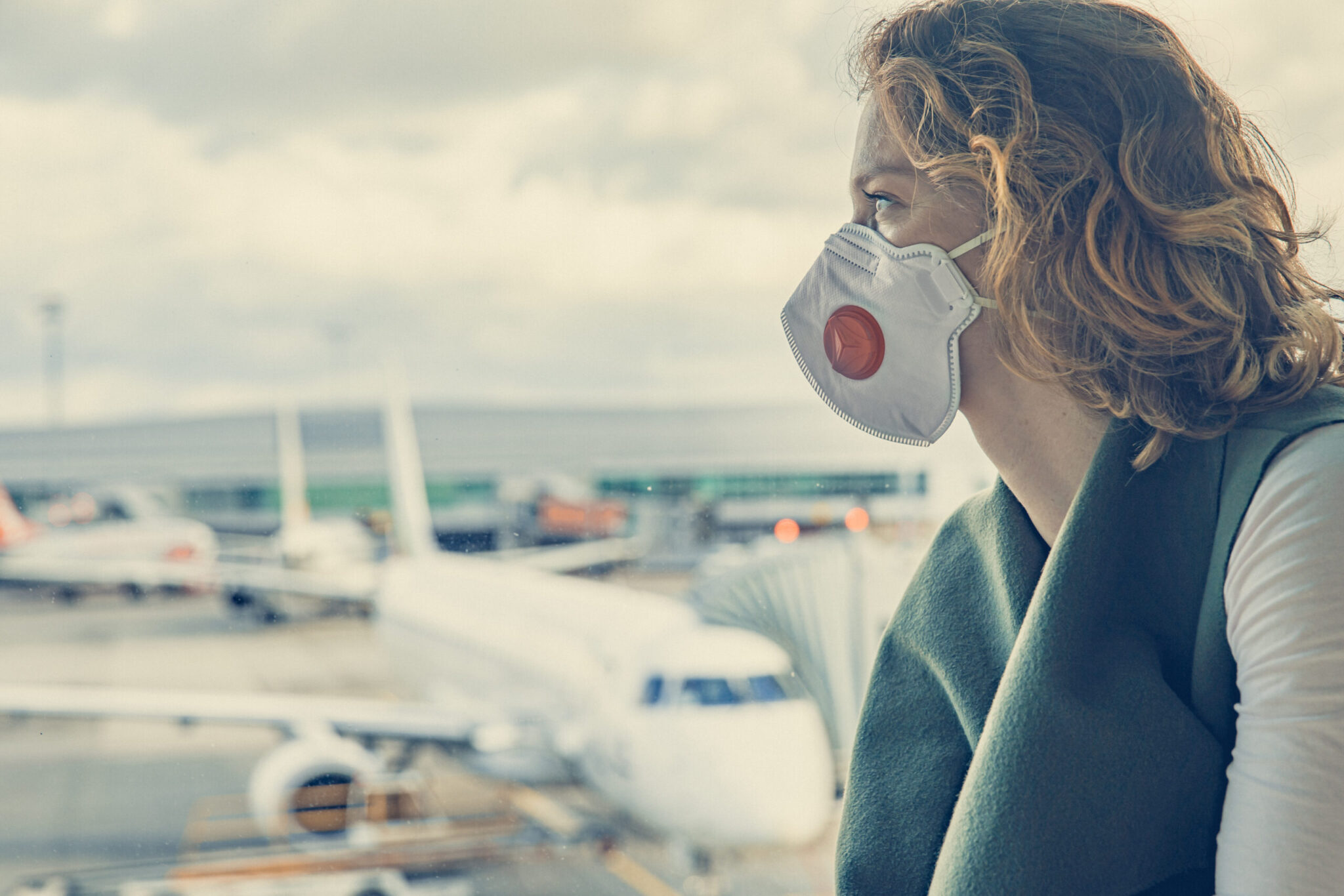 A woman in a medical mask gazes out an airport window at planes on the tarmac, reflecting the themes of 