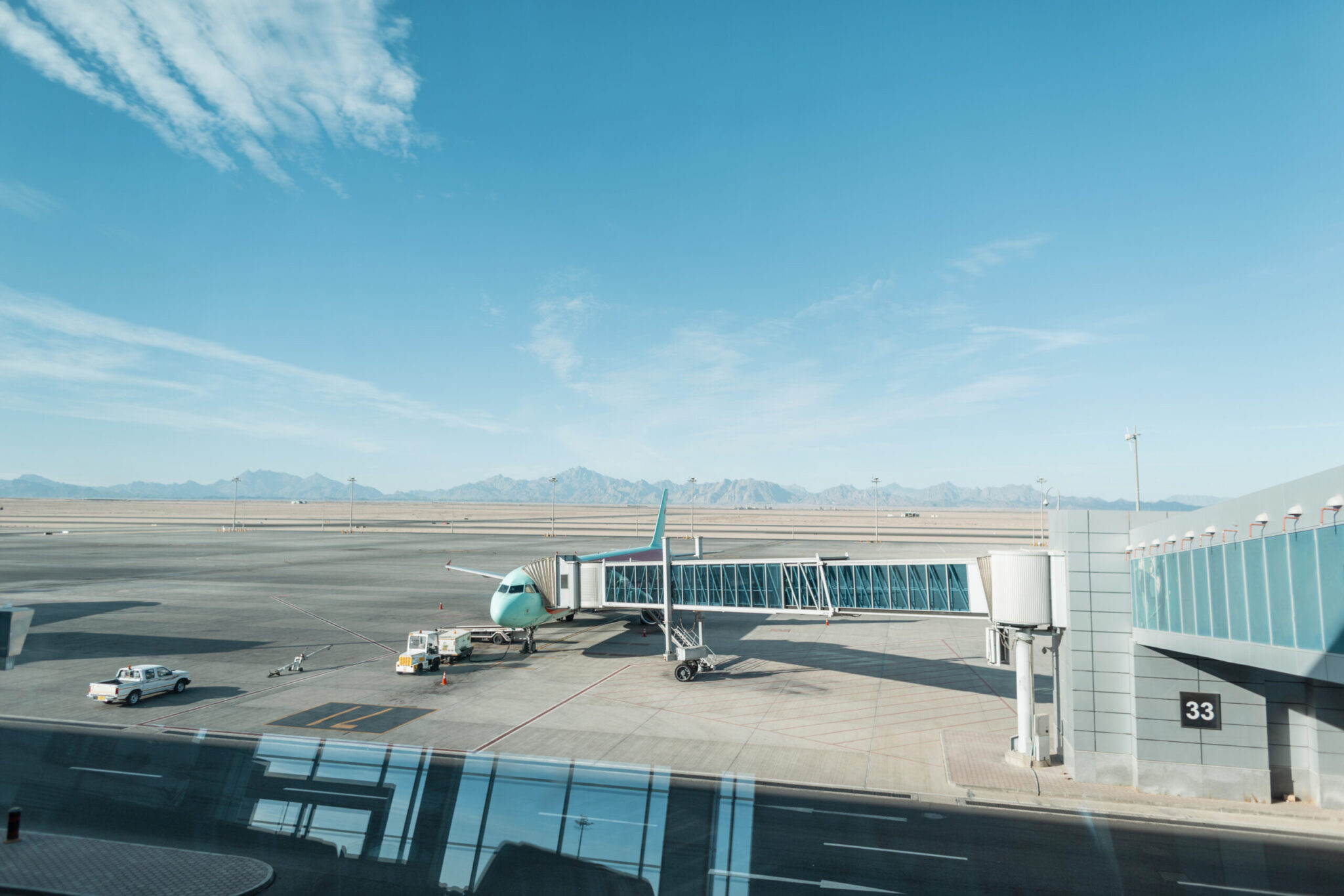 A turquoise and white airplane is parked at an airport gate beneath clear skies and mountains, promoting the 