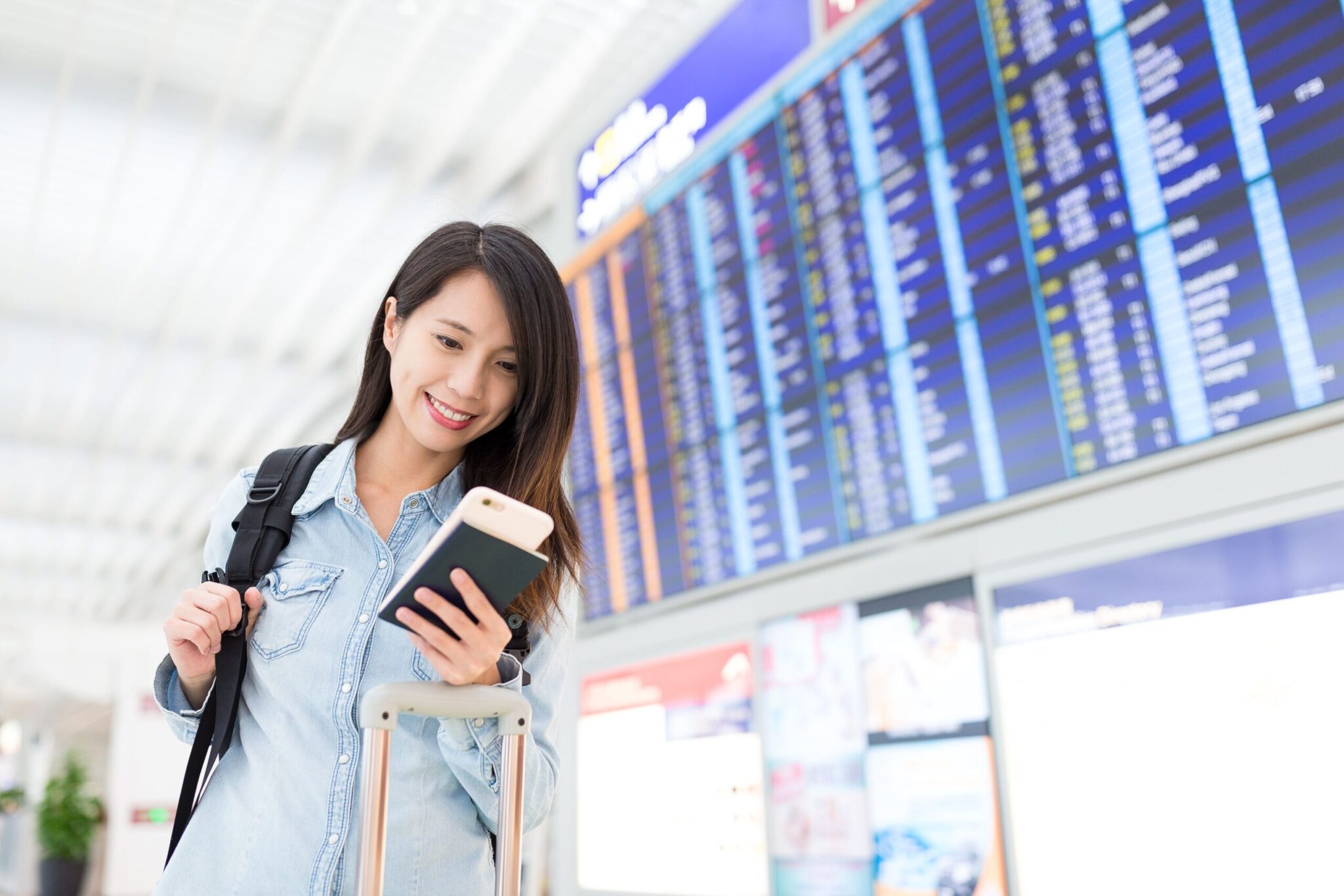 A woman at an airport smiles at her phone, backpack and suitcase nearby, with flight info screens behind her—highlighting themes from 