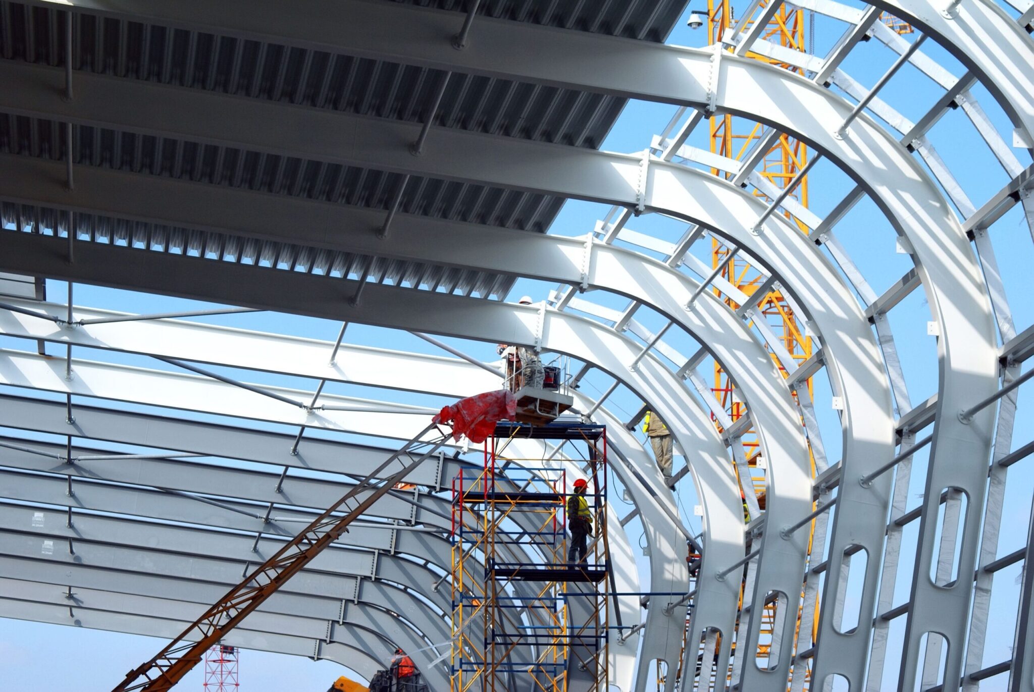 Construction workers on lifts assemble a large, curved metal roof for an airport under blue sky, with cranes and scaffolding—a scene reflecting trends in the Global Outlook of Airport Capital Expenditure.