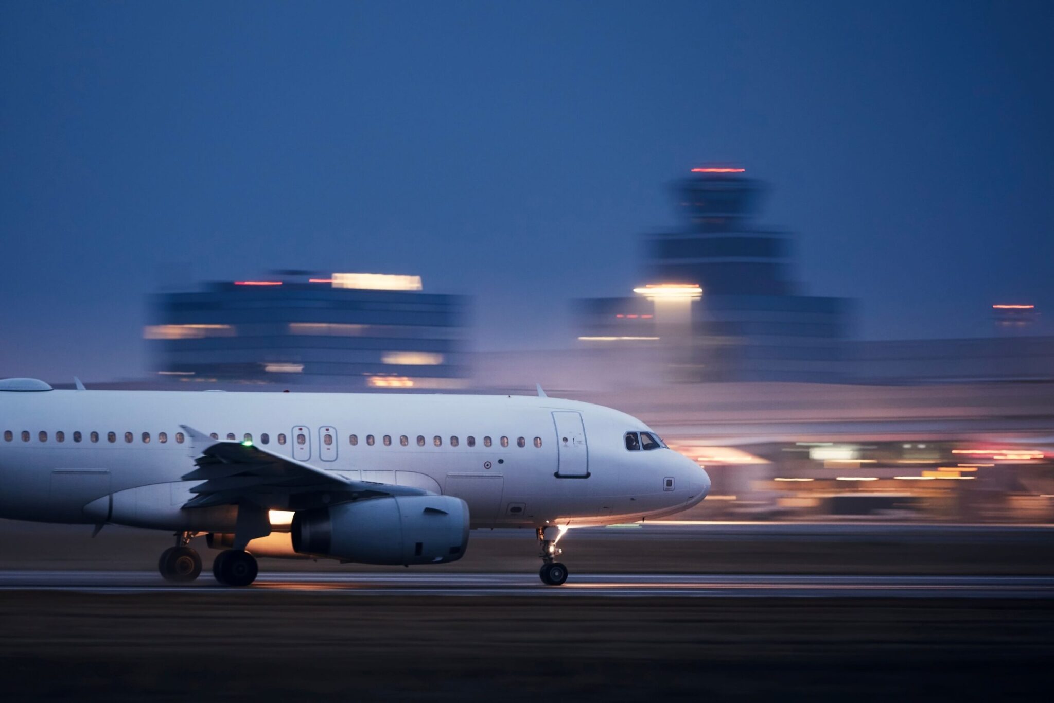 A white commercial airplane with its lights on approaches the runway at dusk, airport buildings aglow in the background, symbolizing the safe arrivals enabled by the One-Stop Security Toolkit.