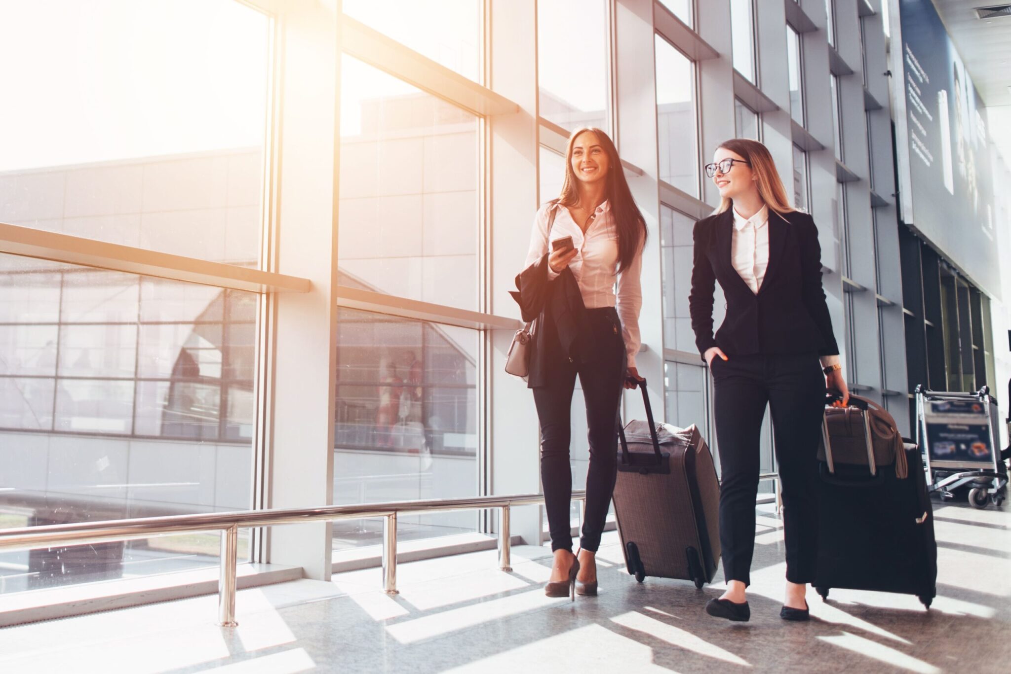 Two women in business attire walk through a bright, modern airport terminal with suitcases, reflecting the efficient travel experience outlined in the Passenger Facilitation Performance Metrics Handbook (2020).