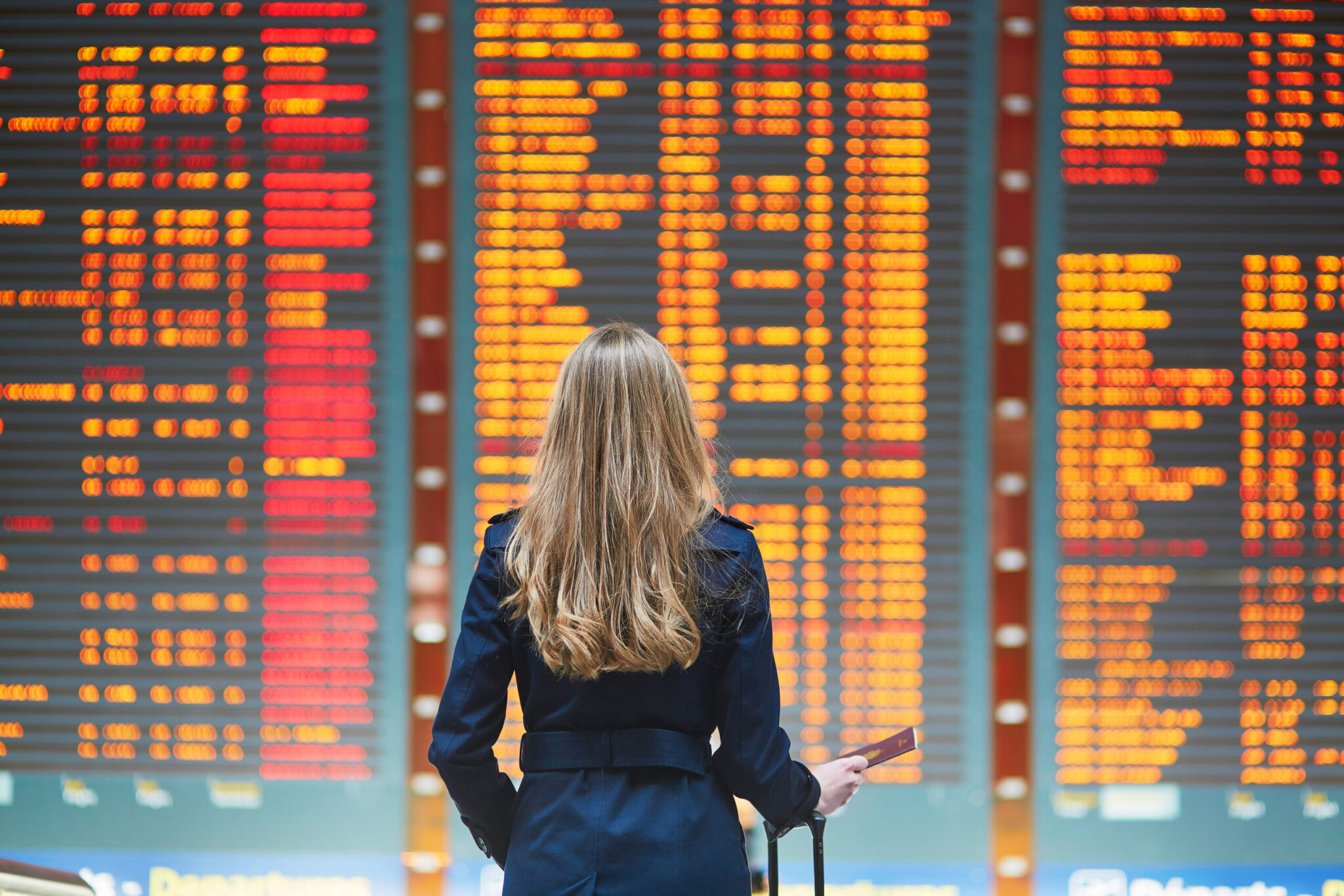 A woman with long blonde hair, wearing a dark coat and holding a suitcase, reviews flight boards at the airport—highlighting themes from 
