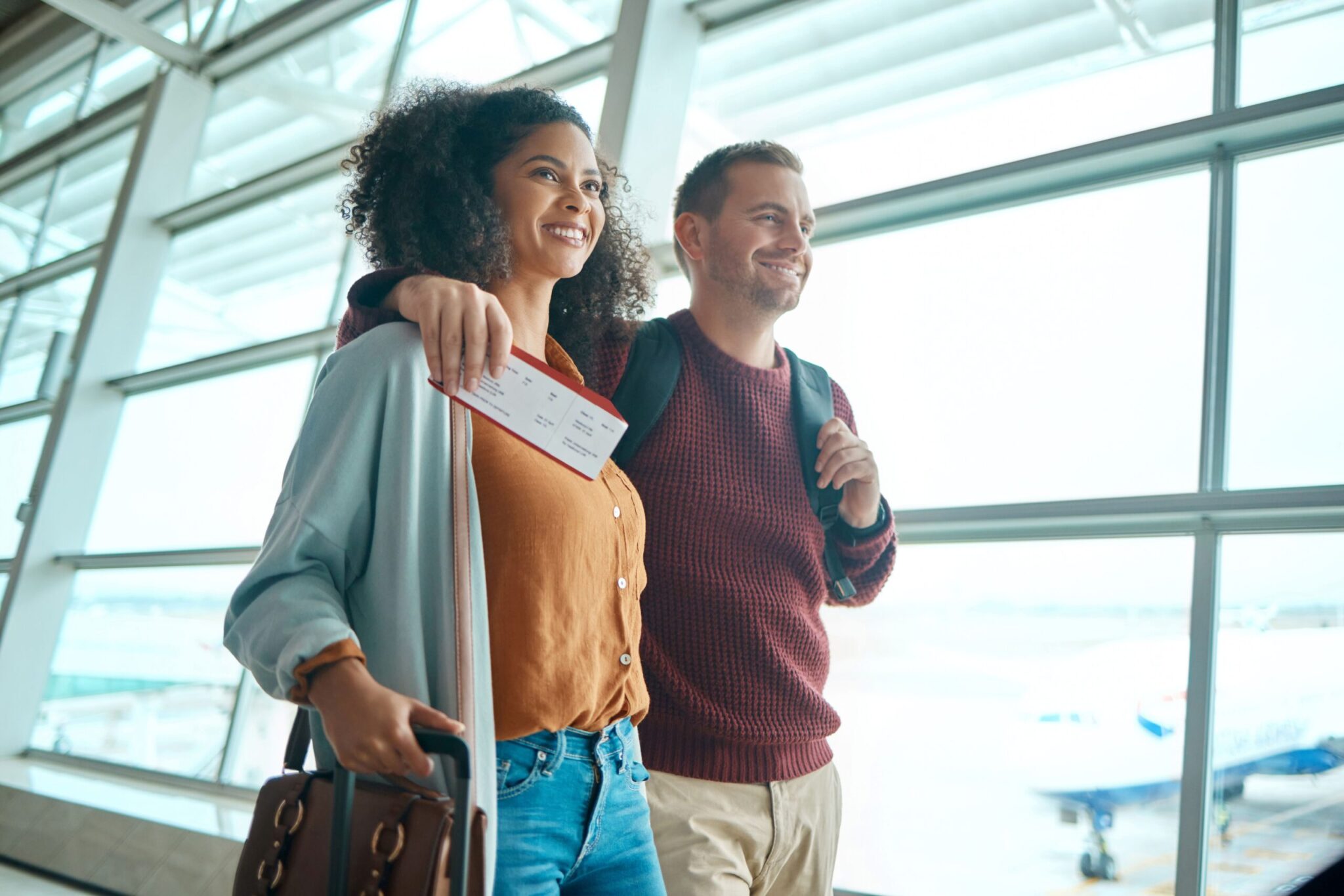 A couple walks through an airport, smiling as the woman holds a boarding pass and purse while the man carries a backpack. This scene captures elements explored in 