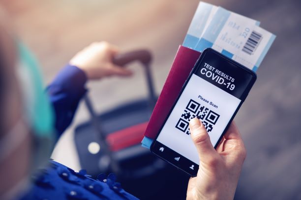 A person holds a smartphone showing a COVID-19 test result QR code, airline tickets, and a passport while pulling a suitcase, illustrating 