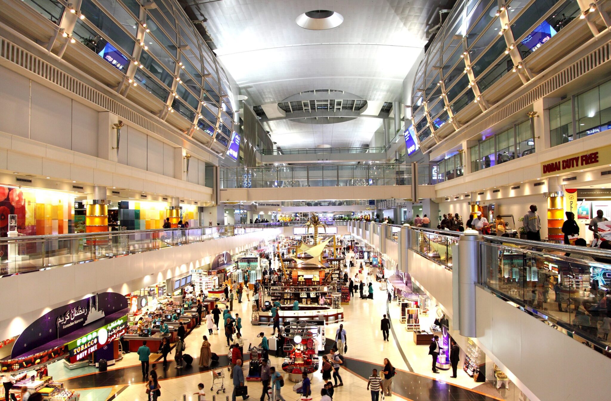 A bustling airport terminal features modern architecture, glass railings, bright lighting, and a duty-free shop. Discover insights on retail spaces in our White Paper: Airport Concession Agreements.