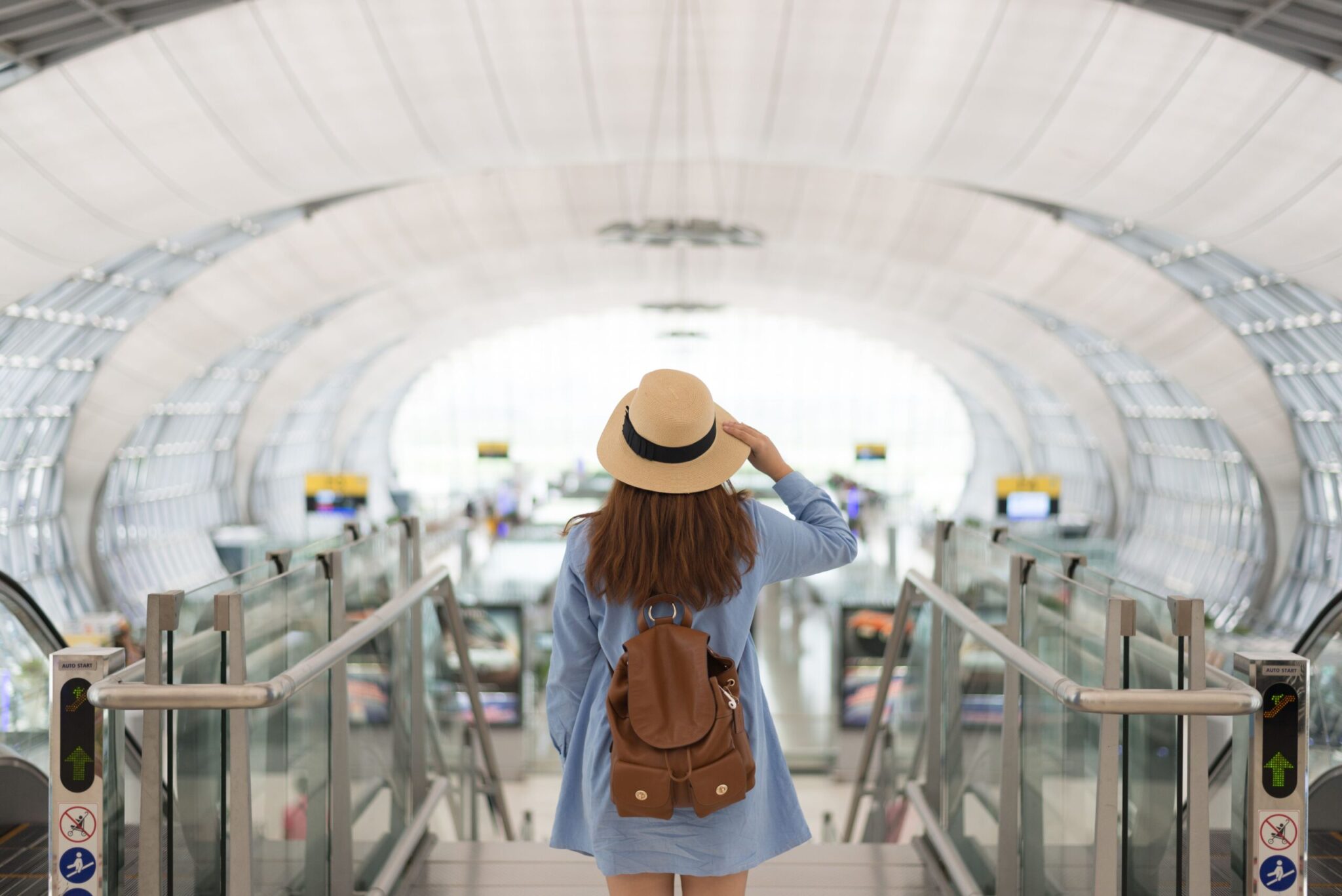 A woman with long hair, wearing a hat, blue jacket, and brown backpack stands at the top of an airport escalator, gazing toward bright windows—an image captured for the White Paper - Path to Recovery: The Airport Retail Perspective.