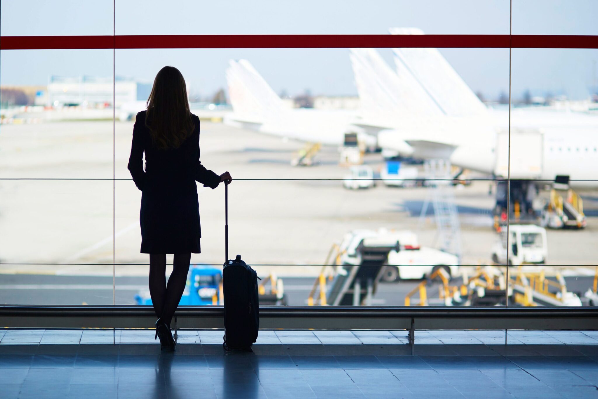 At an airport terminal, a woman with long hair holds her suitcase and gazes out the window at planes, reflecting themes from the ACI Annual Report 2020.
