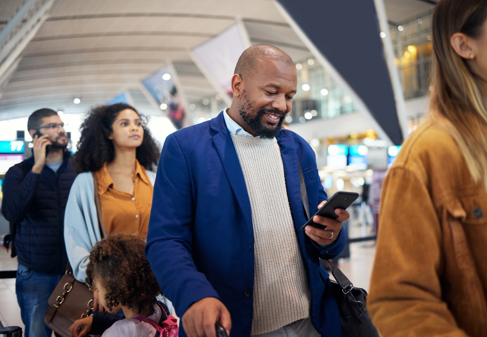 A man in a blue blazer smiles at his phone while standing with other travelers in a busy, bright airport terminal—illustrating the need for tools like the Webinar: Sharing Airport Wait Times – The Opportunities of Data Exchange.