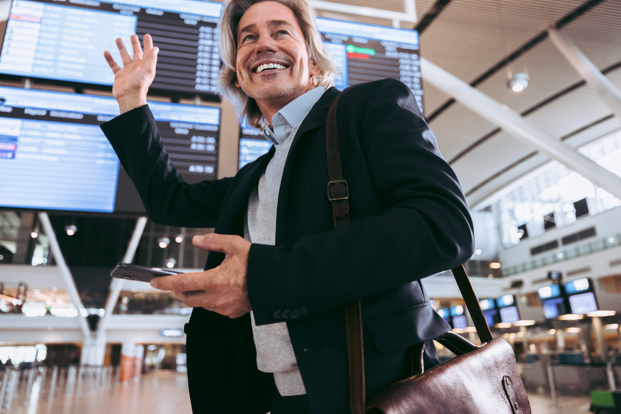 A businessman with a leather bag checks his smartphone in an airport terminal, flight info boards behind him, referencing the ACI World Airport Traffic Forecasts 2023–2052.