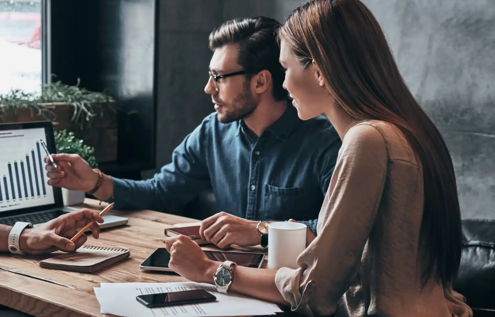 Two people sit at a wooden table with papers, a notebook, a phone, a cup, and pen as they review bar graphs on a laptop using the Intelligence Hub - Analyst Module (Monthly Refreshed Data) and discuss insights together.