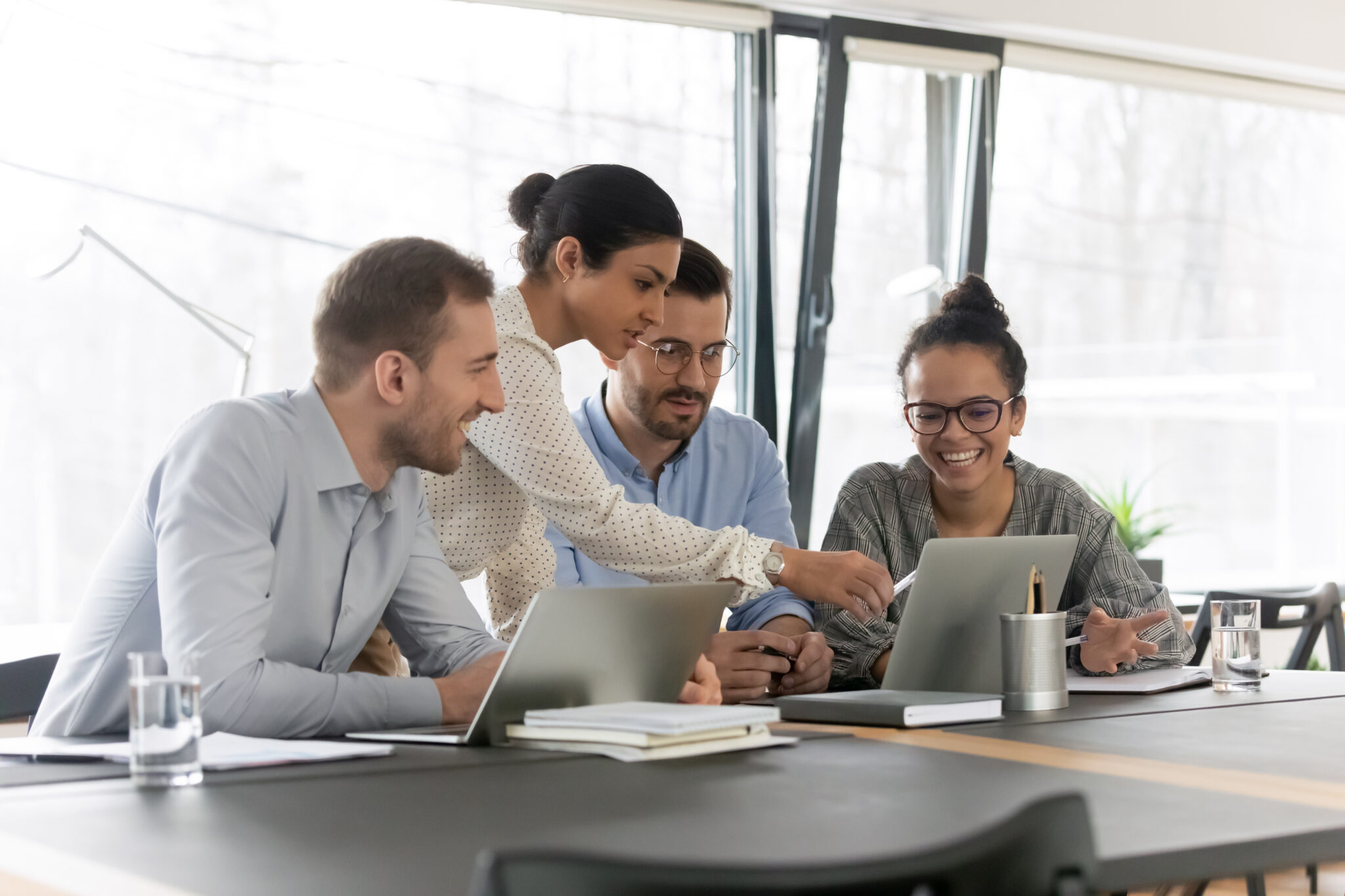 Four colleagues collaborate around a table with laptops and notebooks, smiling as one woman points to another’s screen while using the Intelligence Hub - Analyst Module (Historical Dataset) in a bright, modern office.
