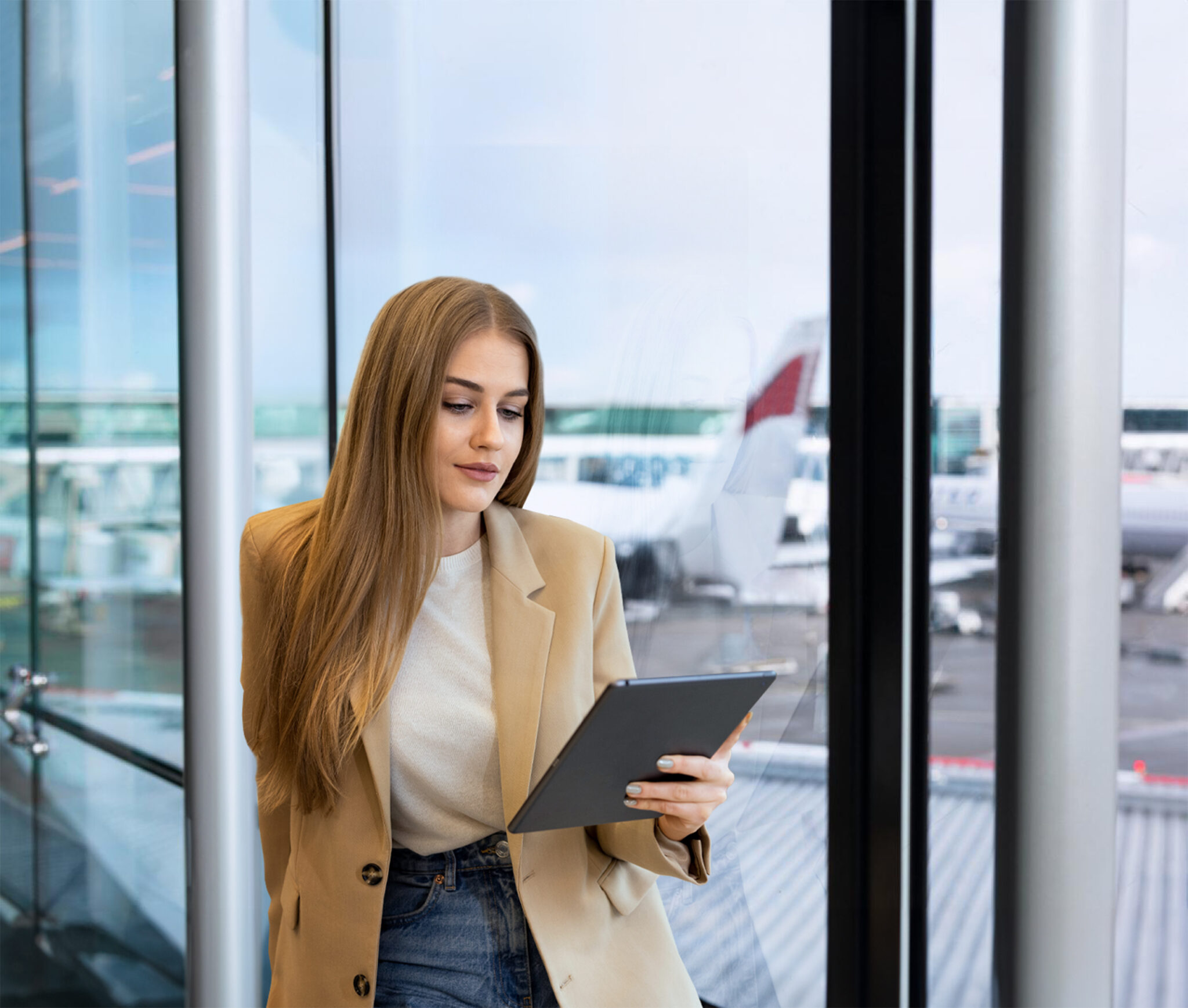 A woman with long blonde hair in a beige blazer and jeans stands by airport windows, looking at a tablet displaying 