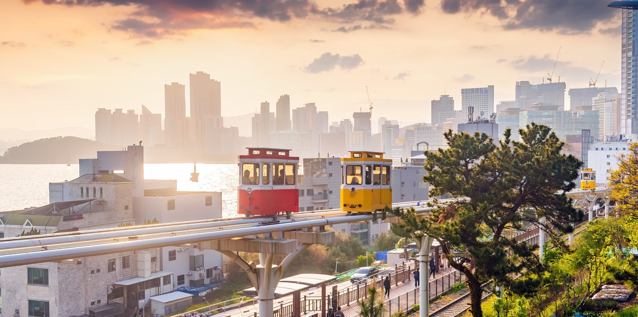 A red and a yellow tram travel on elevated tracks above a cityscape at sunset in the Airports Innovate 2025 scene, with high-rise buildings, water, and trees in the background.