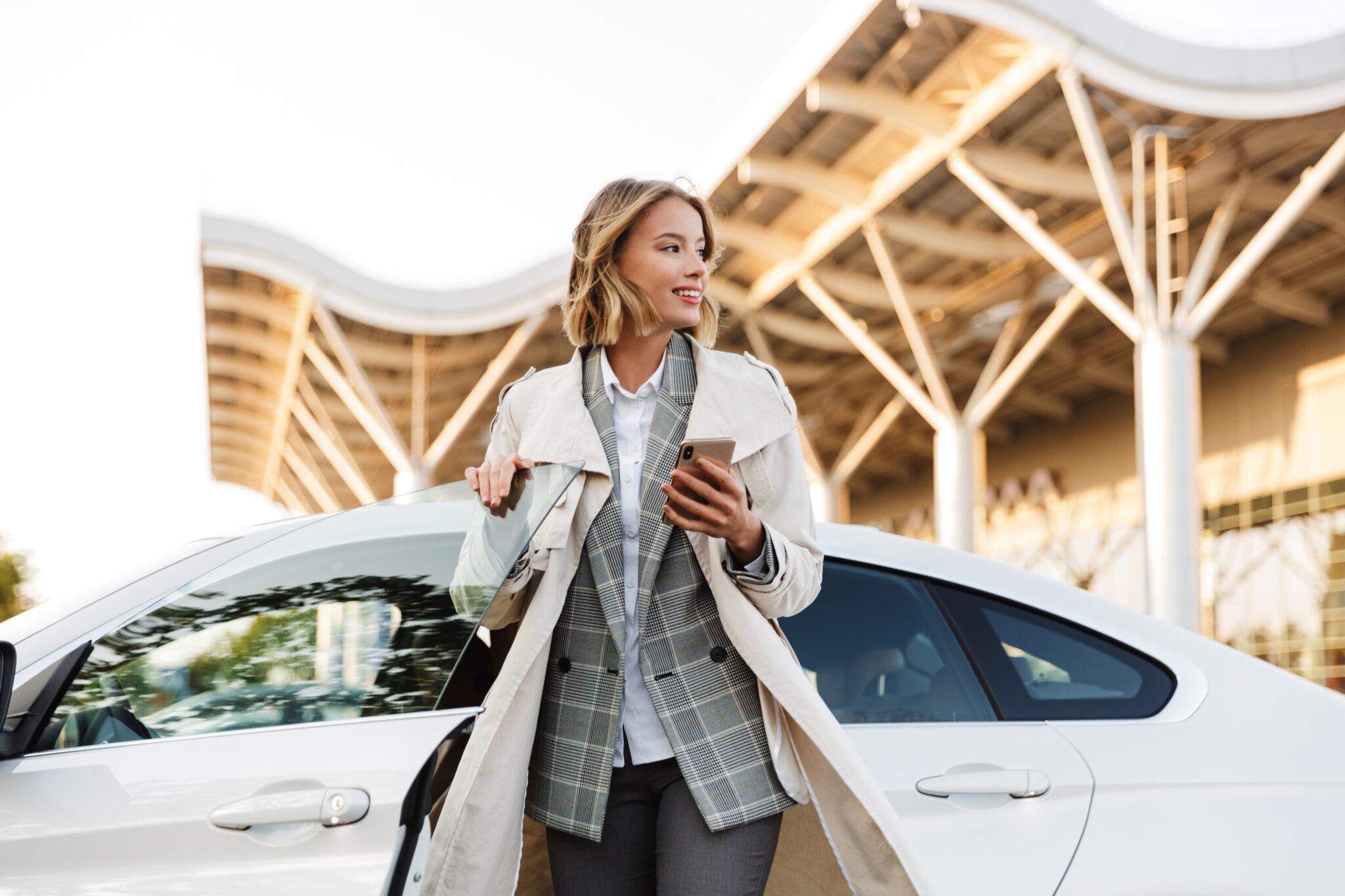 A woman in a light trench coat stands by a white car, smiling and holding a smartphone, with modern architecture behind her—featured on the cover of the 
