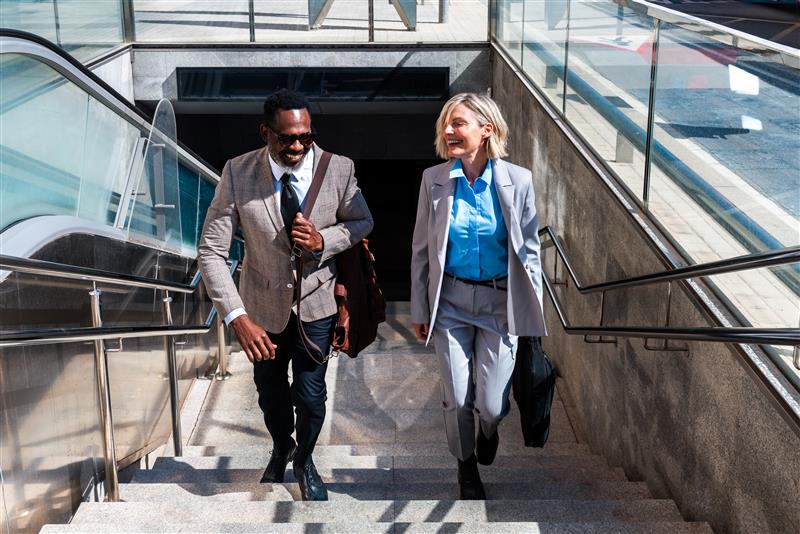 Two business professionals in suits walk up sunlit outdoor stairs, smiling and carrying bags, with modern glass and stone architecture—a perfect setting for the 2025 Airport Workforce Management Report.
