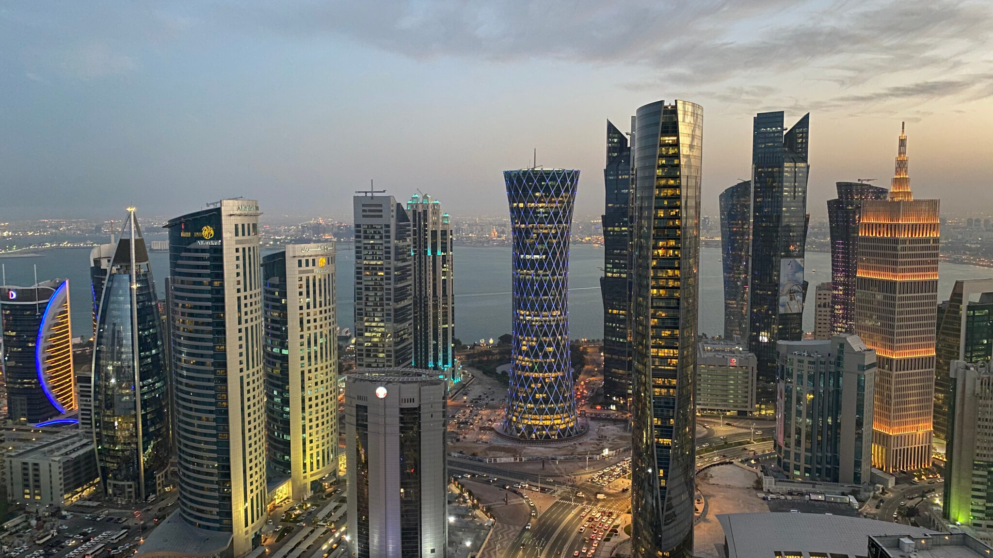 The Trinity Forum depicts a cityscape at dusk with tall, modern skyscrapers—some lit up—surrounding a curved bay, partly cloudy skies above and city lights reflecting on the water in the background.