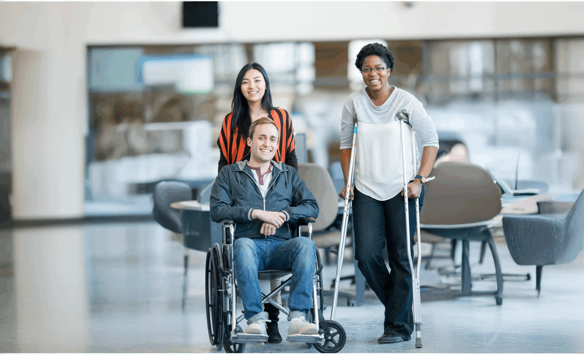 Three smiling people—one man in a wheelchair, a woman behind him, and another with crutches—are in a modern office, embodying the inclusivity promoted by 