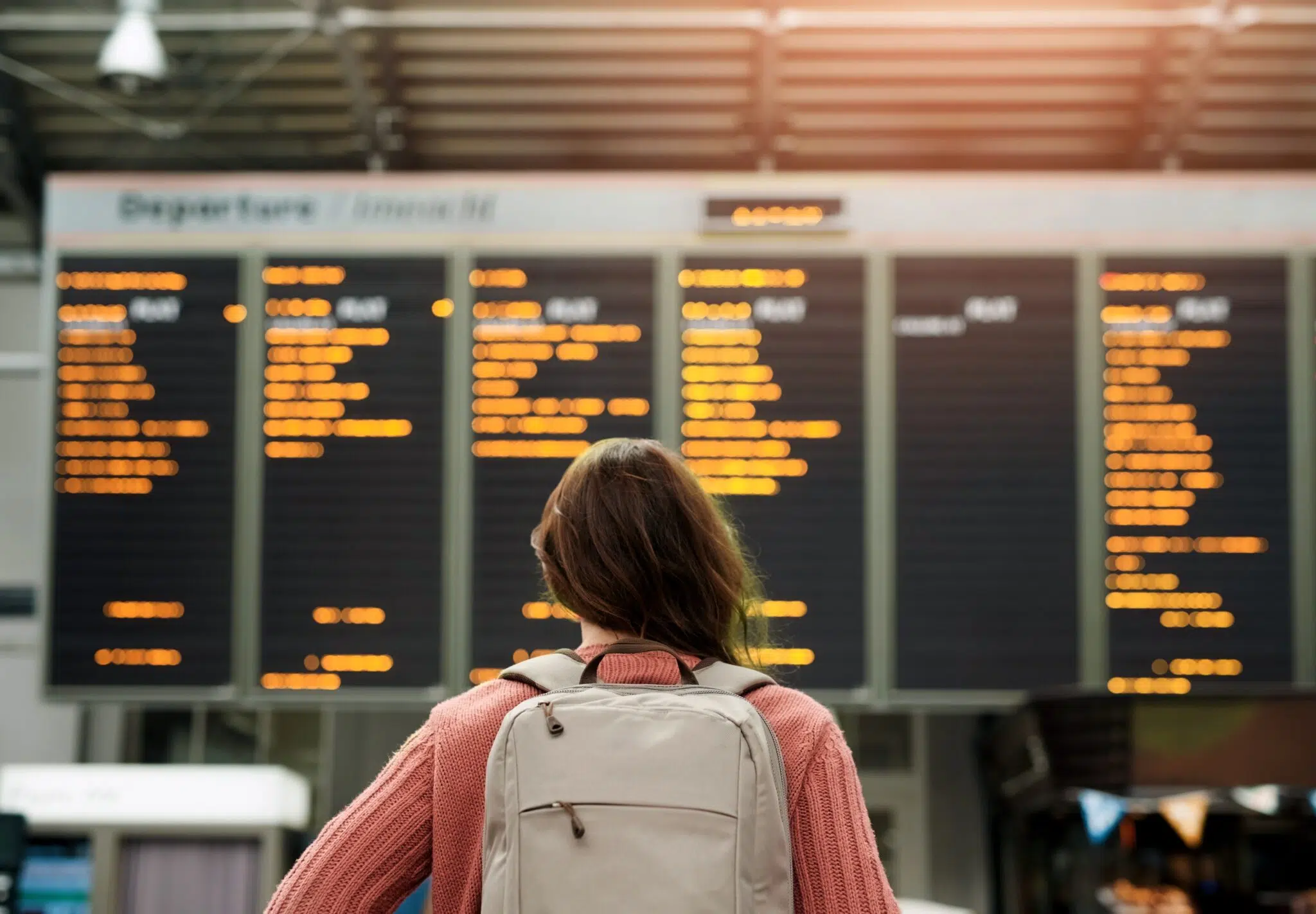 A person with long hair and a backpack stands before a large digital departures board at an airport, checking flight details from the Annual World Airport Traffic Dataset, 2026.